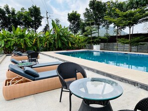 A poolside area with lounge chairs and a glass table, surrounded by lush greenery and a view of a mountain. - Echoes Retreat (Doddaballapur)
