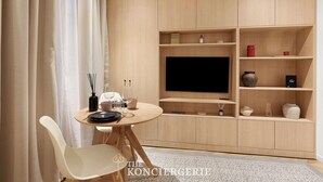 Un salon moderne avec une table ronde en bois, des chaises blanches, un téléviseur à écran plat et des étagères en bois intégrées, agrémentées d'objets décoratifs.