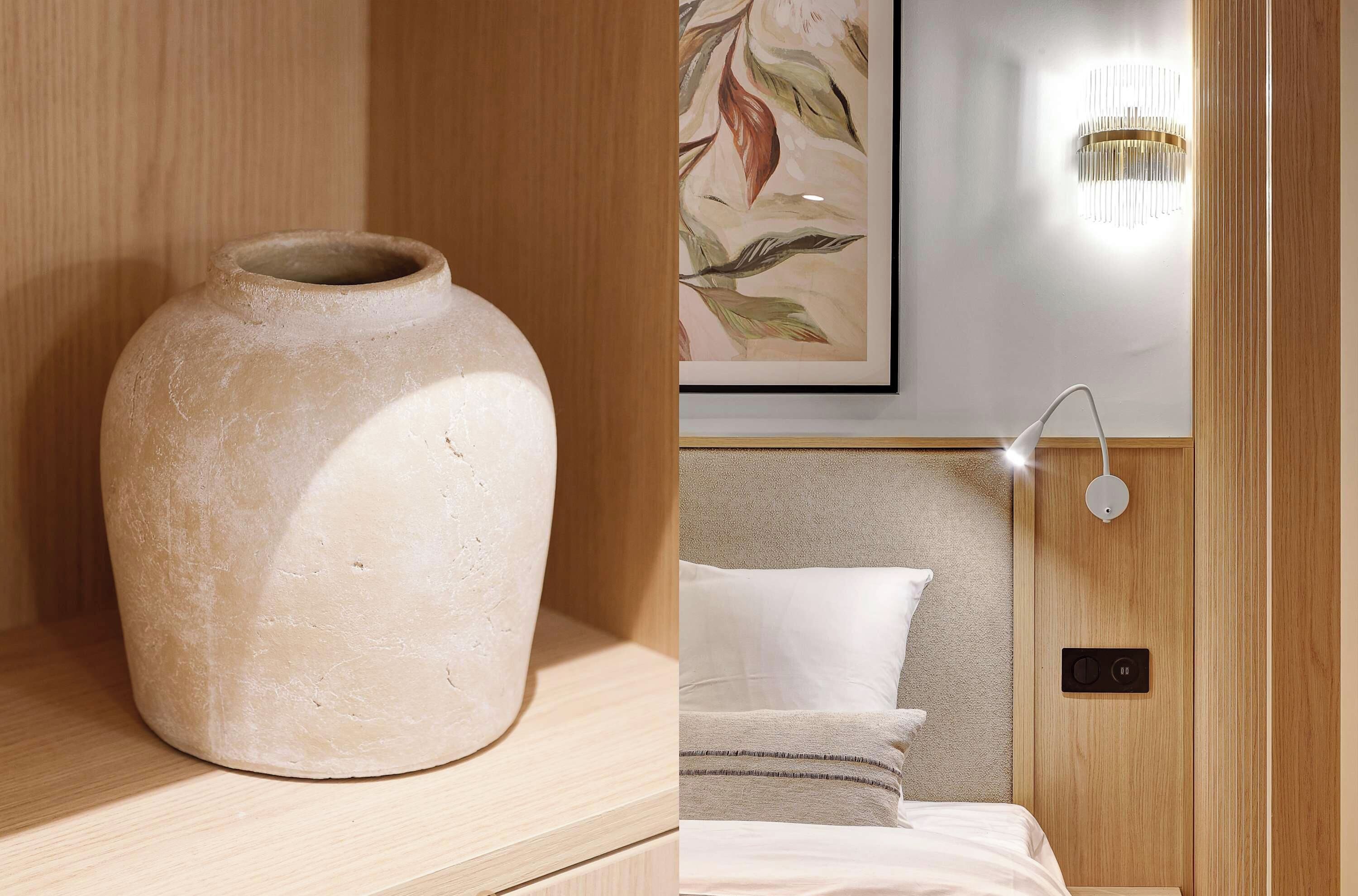 A room with a large vase on a wooden shelf, a bed with a headboard, and a wall-mounted light fixture.