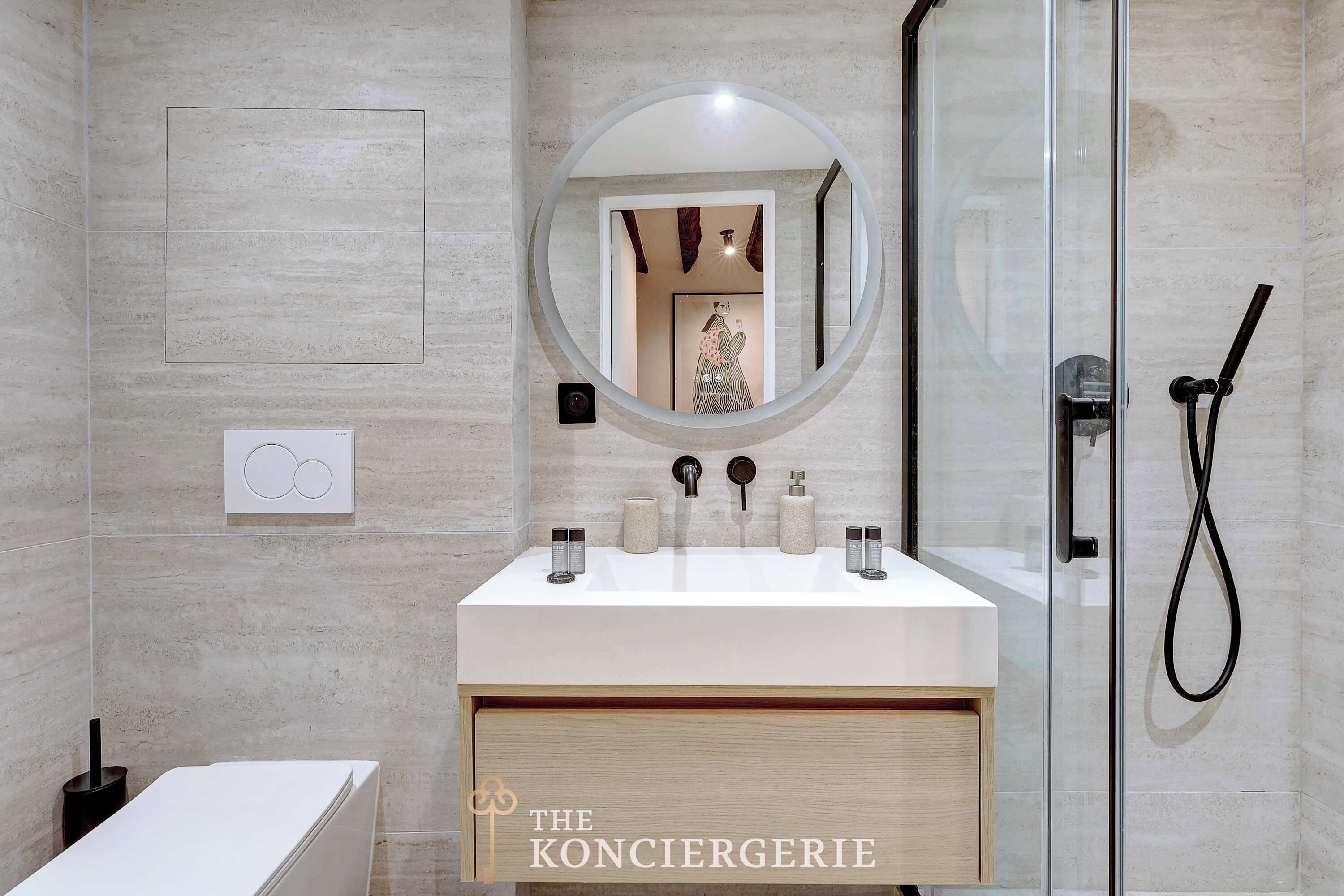 A modern bathroom with a glass shower enclosure, a round mirror, and a white sink with two faucets.