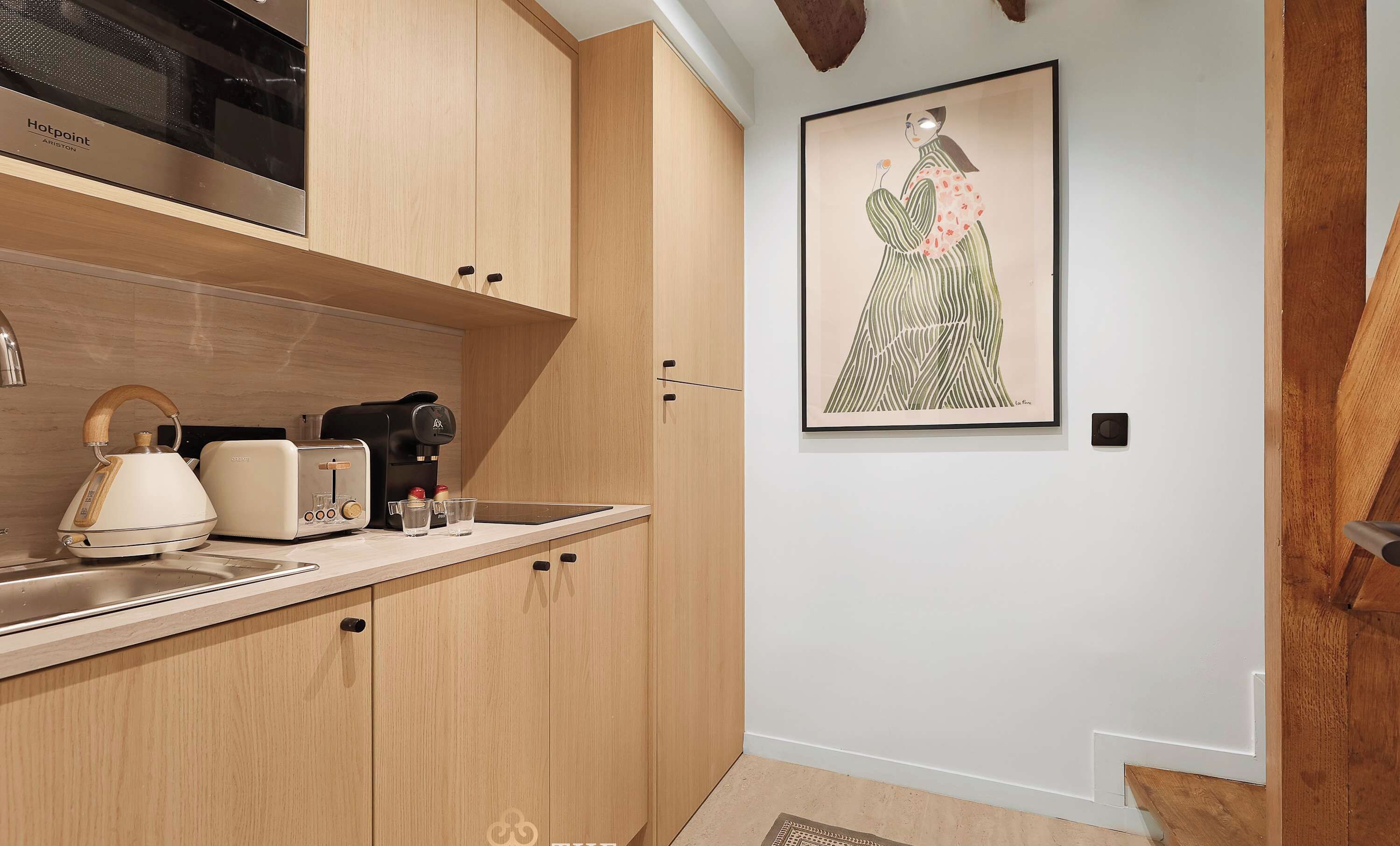 Une cuisine moderne avec des meubles en bois, un four à micro-ondes, une bouilloire et un grille-pain. Un tableau encadré orne le mur et le sol est recouvert de parquet.