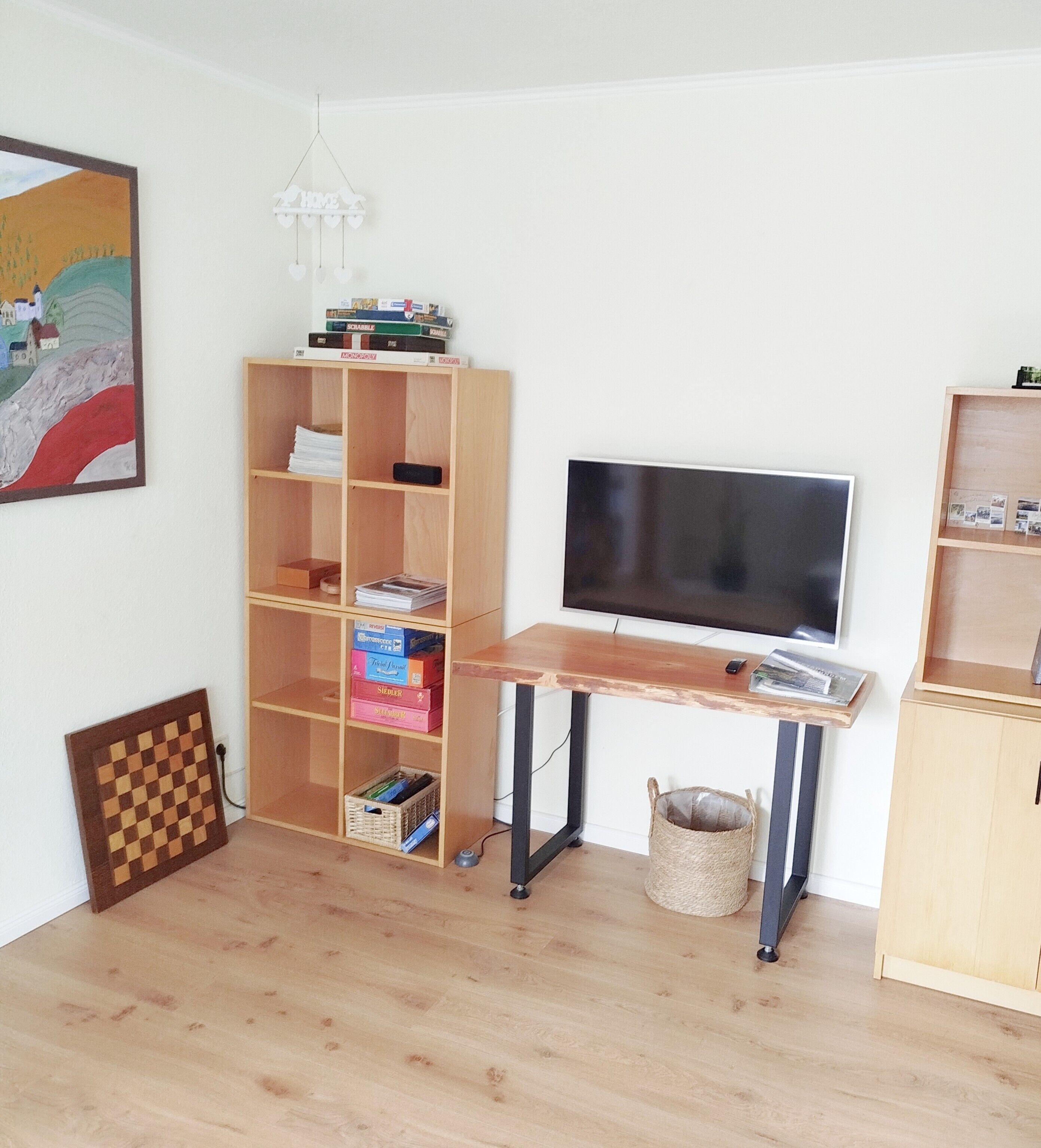 Una habitación con piso de madera, televisor sobre un mueble, una estantería y un cuadro en la pared.