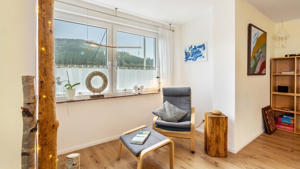 Ein Zimmer mit großem Fenster, einem Sessel, einem kleinen Tisch und einem Bücherregal.
