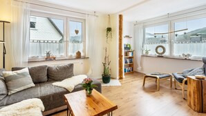 Ein gemütliches Wohnzimmer mit Sofa, Couchtisch und Sesseln, einem Bücherregal und einem großen Fenster mit Blick auf eine verschneite Landschaft.