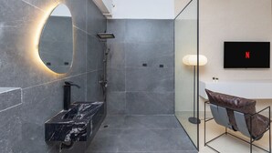 Un baño moderno con lavamanos de mármol, cabina de ducha con mampara de vidrio y un televisor con el logo de Netflix.