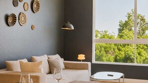 Ein modernes Wohnzimmer mit einer Couch, einem Esstisch für zwei Personen, einer Lampe und Blick auf Bäume durch ein großes Fenster.