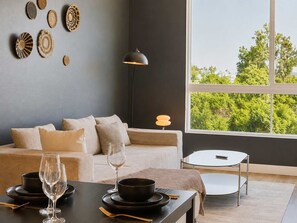 Ein modernes Wohnzimmer mit einer Couch, einem Esstisch für zwei Personen, einer Lampe und Blick auf Bäume durch ein großes Fenster.