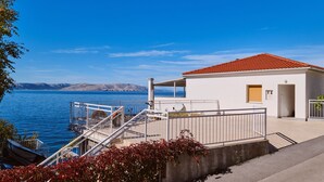Un edificio costero con techo de tejas rojas, una escalera que lleva a un balcón y vistas al mar y montañas lejanas.