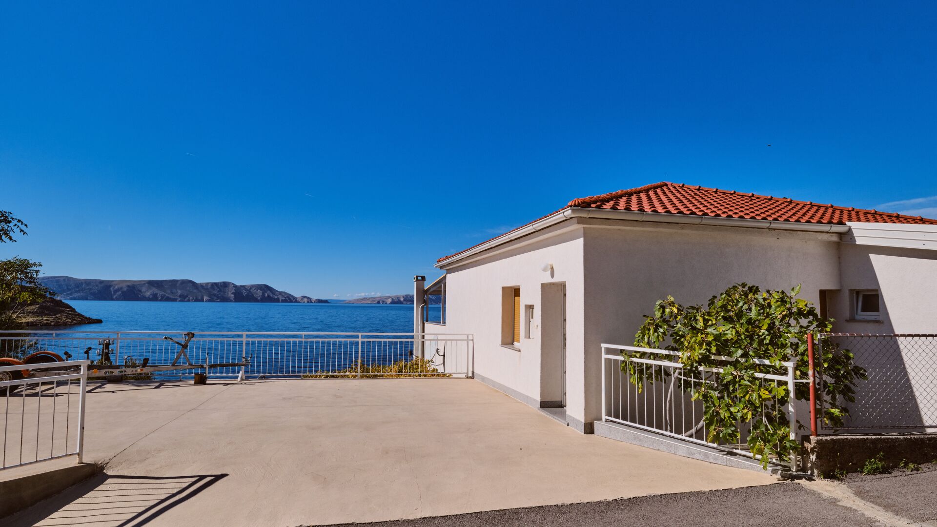 A coastal building with a red-tiled roof, a concrete patio, and a view of the sea and distant mountains.