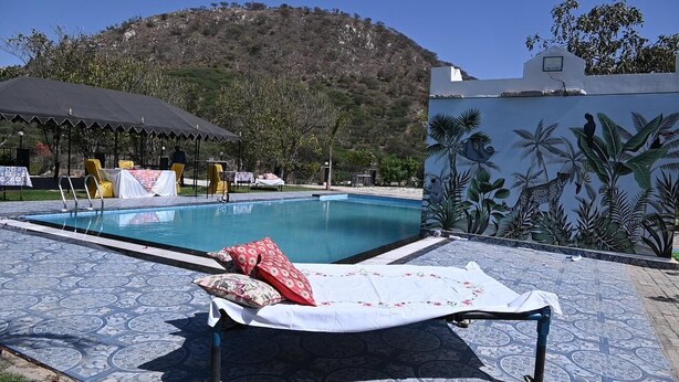 A pool area with a lounge chair, a covered seating area, and a mural of tropical plants. - JHAR ARRAVLI RESORT  (Bassi)