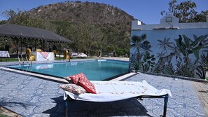 A pool area with a lounge chair, a covered seating area, and a mural of tropical plants. - JHAR ARRAVLI RESORT  (Bassi)
