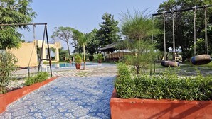 A pool area with a swing set, potted plants, and a tiled walkway. - JHAR ARRAVLI RESORT  (Bassi)