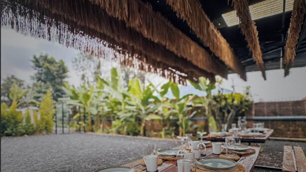 Un área de comedor al aire libre con techo de paja y mesas de madera dispuestas para una comida, rodeada de abundante vegetación.