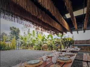 An outdoor dining area with thatched ceiling and wooden tables set for a meal, surrounded by lush greenery. - Pili House & Garden Lodge Rwanda (Muhoza)