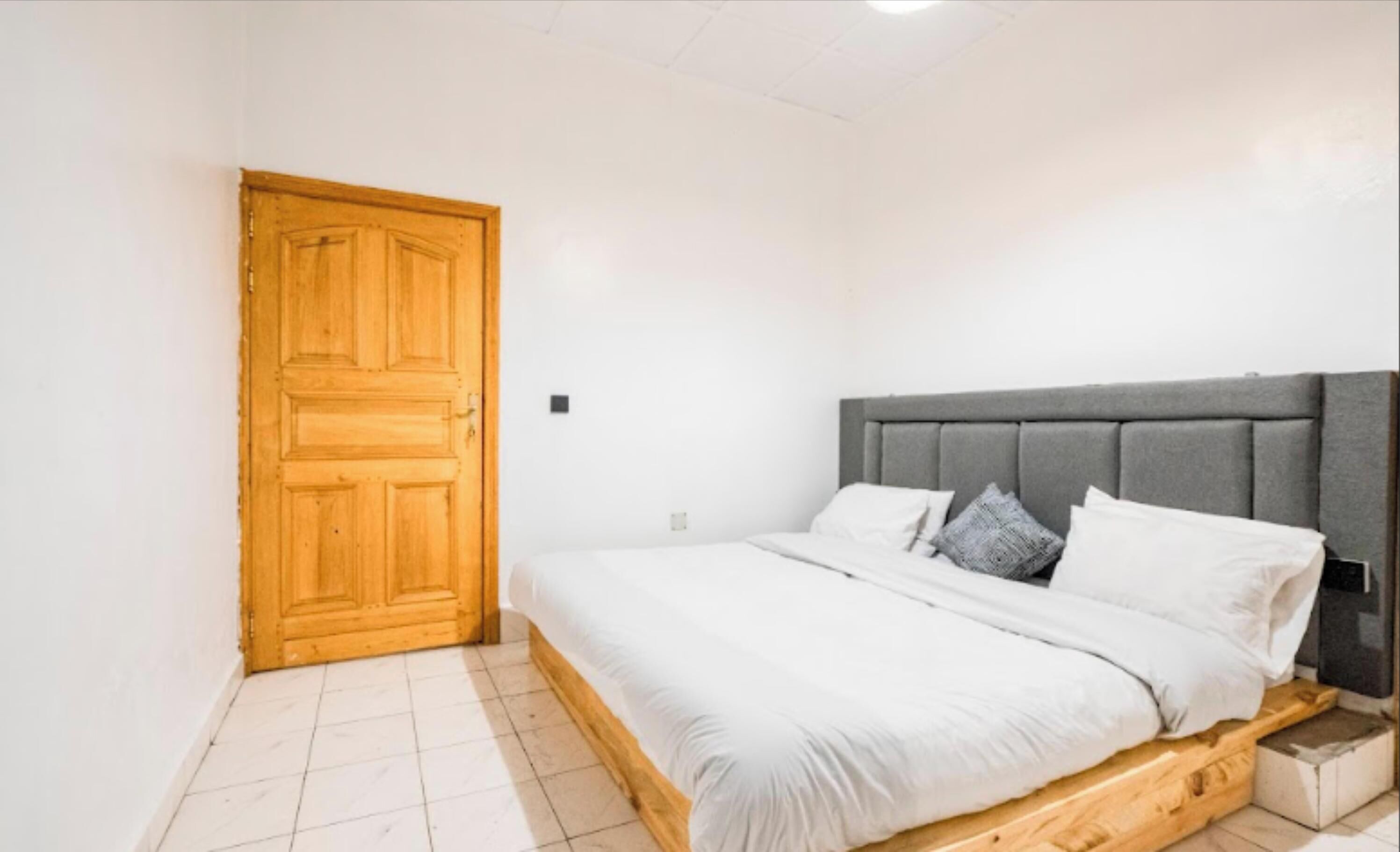 A bedroom with a wooden door, a bed with a grey upholstered headboard, white bedding, and a small wooden nightstand.