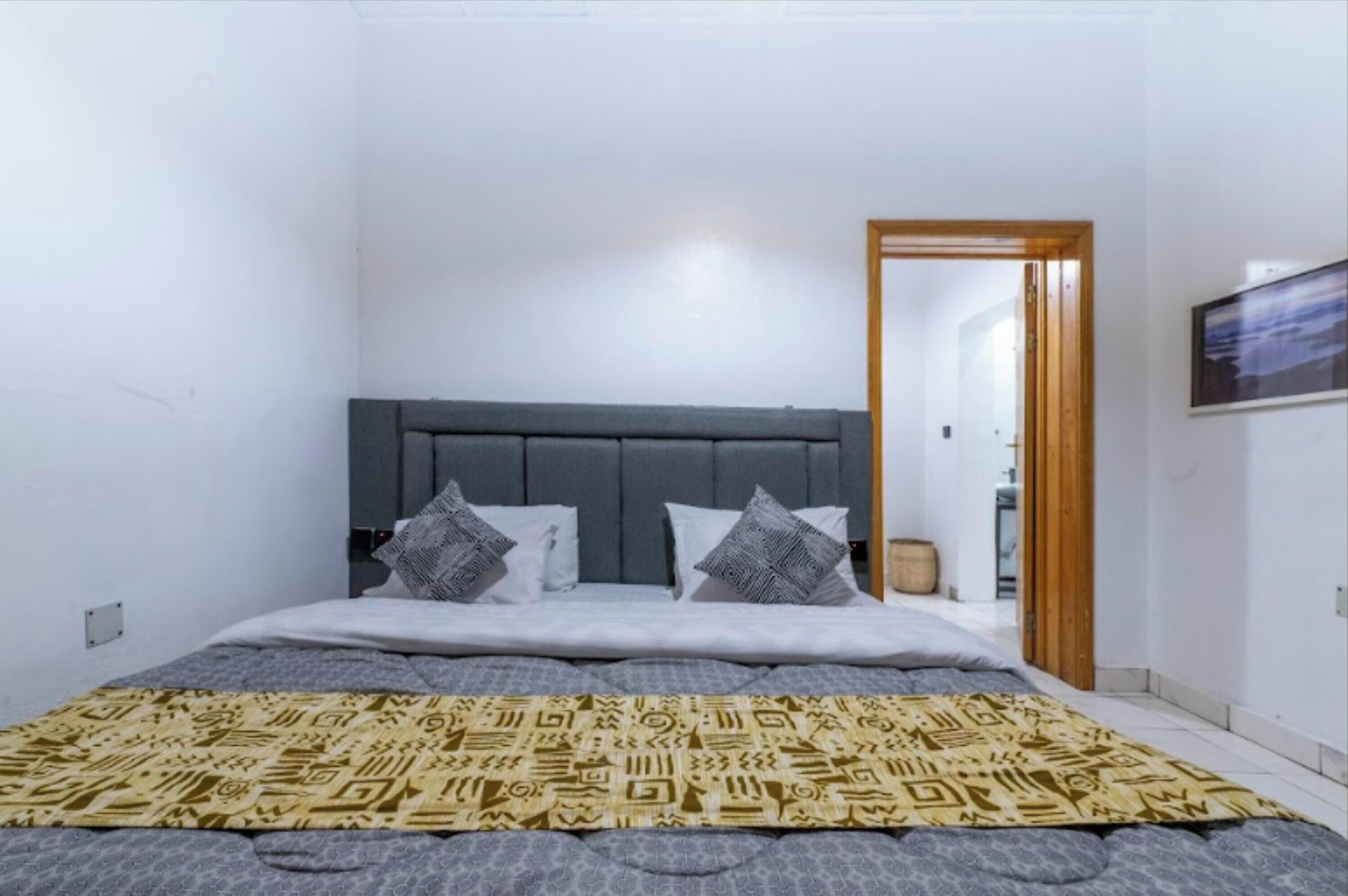Un dormitorio con cabecera gris, edredón estampado y puerta de madera que da a otra habitación.