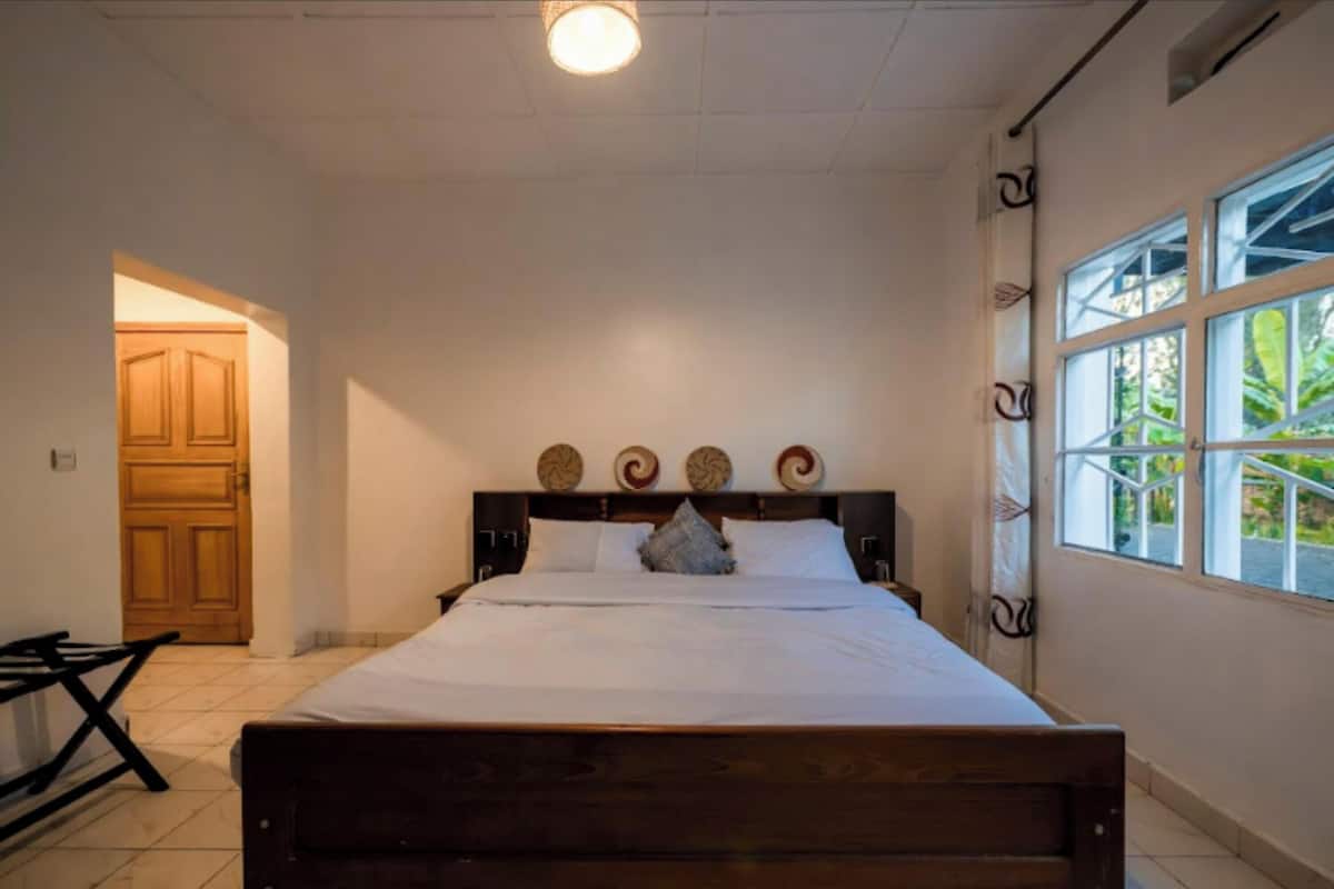 A bedroom with a wooden bed, a window with a view of greenery, and a door leading to another room.