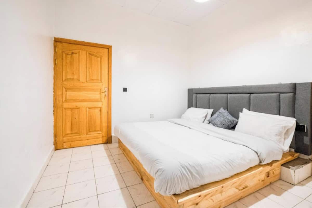 A bedroom with a wooden door, a bed with a grey upholstered headboard, and white bedding.