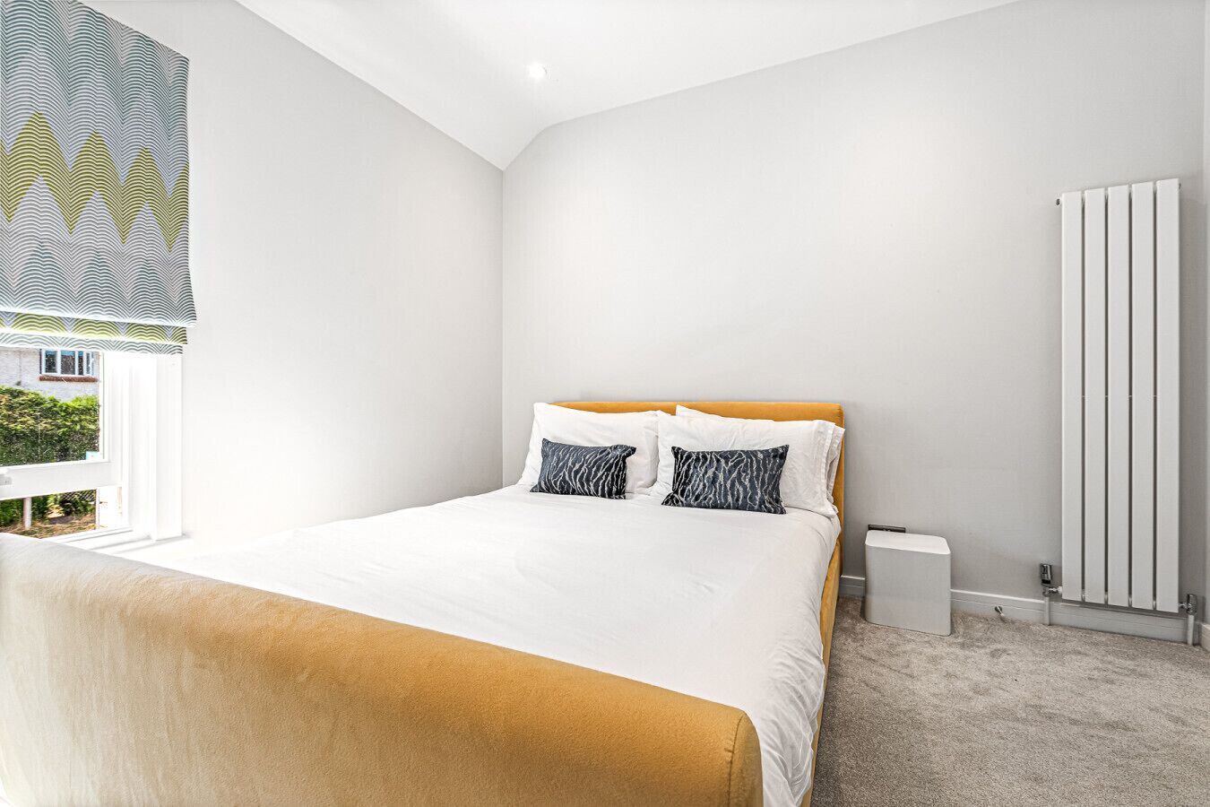 A modern bedroom with a large bed, white walls, a radiator, and a window with a patterned blind.