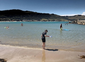 Une plage aux eaux cristallines, où l’on peut voir des gens nager et faire de la planche à voile, avec un paysage vallonné en arrière-plan.