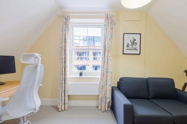 A room with a desk, a chair, a sofa, a window with curtains, and a framed picture on the wall.