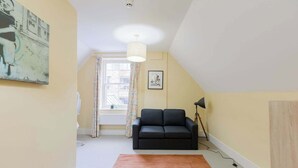 A room with a black sofa, a large window with curtains, a ceiling light, and a framed picture on the wall. - Spacious 3BD Flat in Historic Building Shoreditch (Greater London)