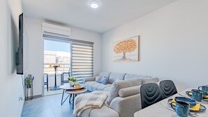 A modern living room with a sofa, a dining table with chairs, a TV mounted on the wall, and a balcony with a view. - TelMar Sky View - “Stylish 3-Bedroom Apartment with Mountain & Sea Views” (Larnaka)