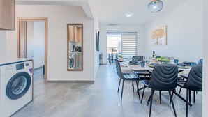 A modern kitchen with wooden cabinets, a built-in washing machine, and a dining area with a table set for four. - TelMar Sky View - “Stylish 3-Bedroom Apartment with Mountain & Sea Views” (Larnaka)