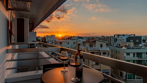 A balcony with a table set for two, two wine glasses, and a bottle of wine. The balcony overlooks a cityscape during sunset. - TelMar Sky View - “Stylish 3-Bedroom Apartment with Mountain & Sea Views” (Larnaka)