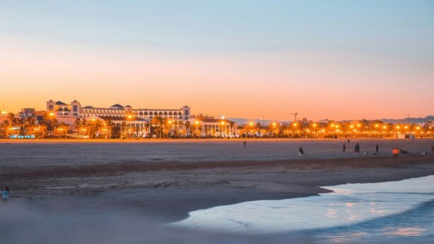 Un hôtel situé en bord de mer, avec une rangée de fenêtres illuminées, une plage de sable et une mer calme.