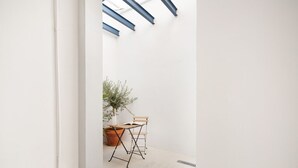 Une pièce minimaliste avec un mur blanc, une grande fenêtre aux poutres bleues, une plante en pot et une table pliante avec deux chaises.