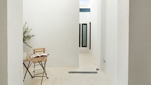 Un couloir étroit avec un mur blanc, un banc en bois sur lequel est posé un livre, et une plante en pot.