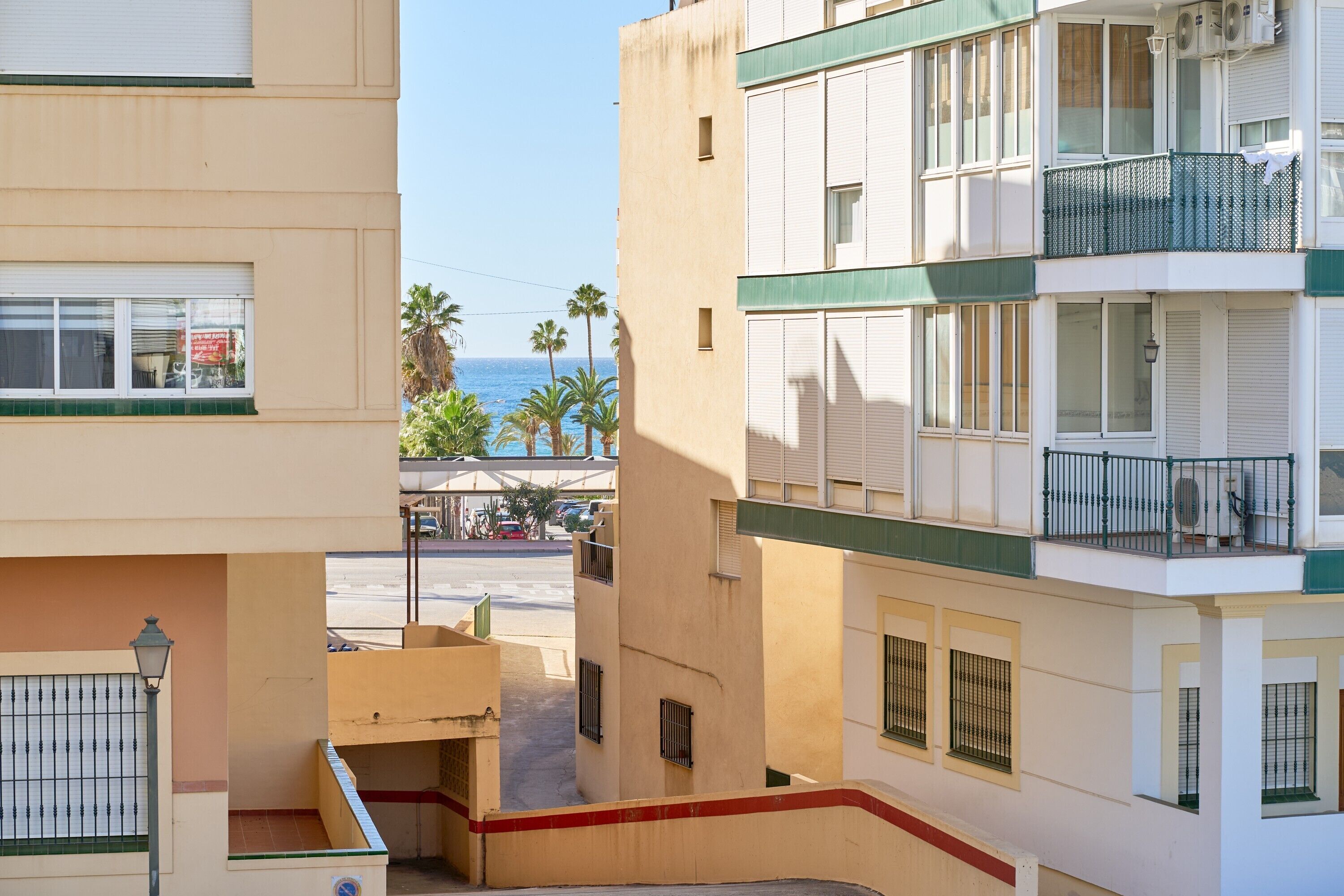 A multi-story building with balconies, a visible street, and palm trees in the background.