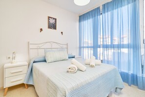 A bedroom with a bed, a nightstand, a lamp, a framed picture, and a window with blue curtains.