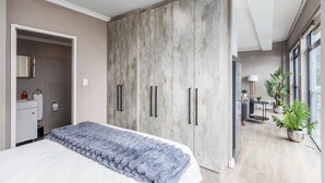 A bedroom with a large wardrobe, a bed with a textured blanket, a bedside table, and a view of a living area with plants and a lamp. - The Square 513 by CTHA (Cape Town)