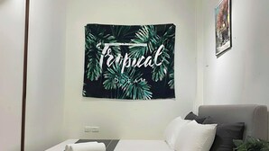 A neatly made bed with a tropical-themed wall hanging, a grey sofa, and a small wooden table with a plant. - NOOK INN OLD QUARTER HANOI (Hanoi)