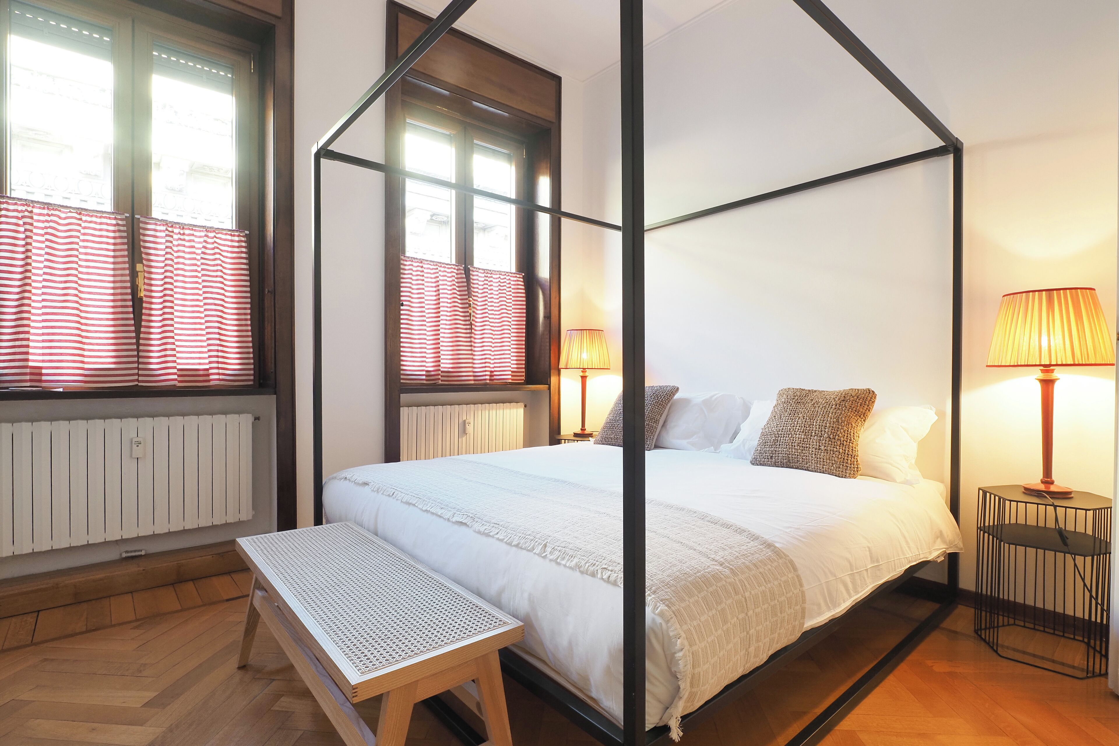 A four-poster bed with a white bedspread and pillows, a wooden bench at the foot of the bed, and two bedside lamps.