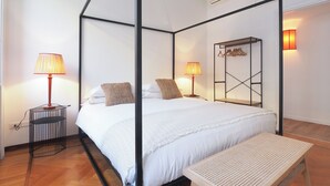 Una cama con dosel y cuatro columnas, con un edredón y almohadas blancos, una mesita de noche con una lámpara y un perchero en la pared.