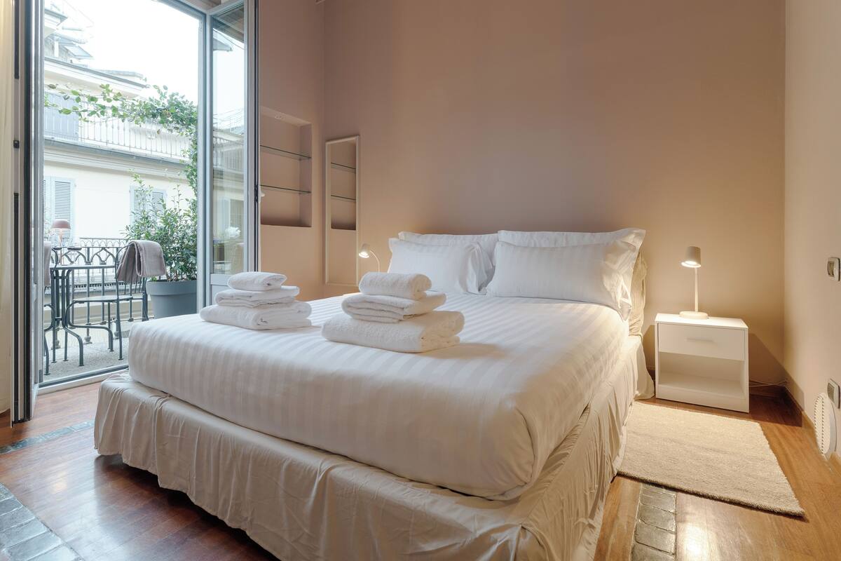 Habitación de hotel con una cama grande, una mesita de noche con lámpara y vistas al balcón.