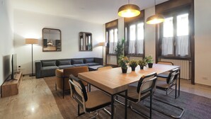 Una sala moderna con una mesa de comedor de madera, asientos cómodos, un televisor de pantalla plana y ventanas grandes con persianas.