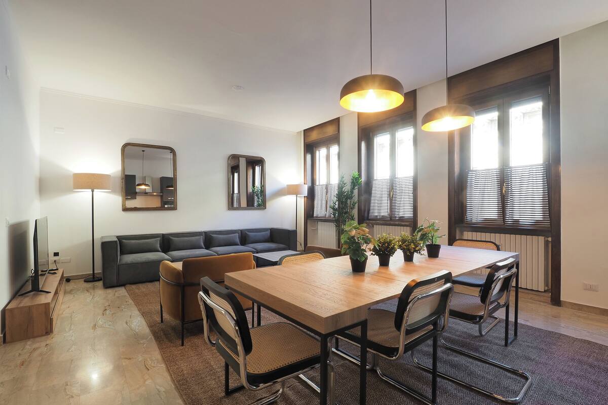 Una sala moderna con una mesa de comedor de madera, asientos cómodos, un televisor de pantalla plana y ventanas grandes con persianas.