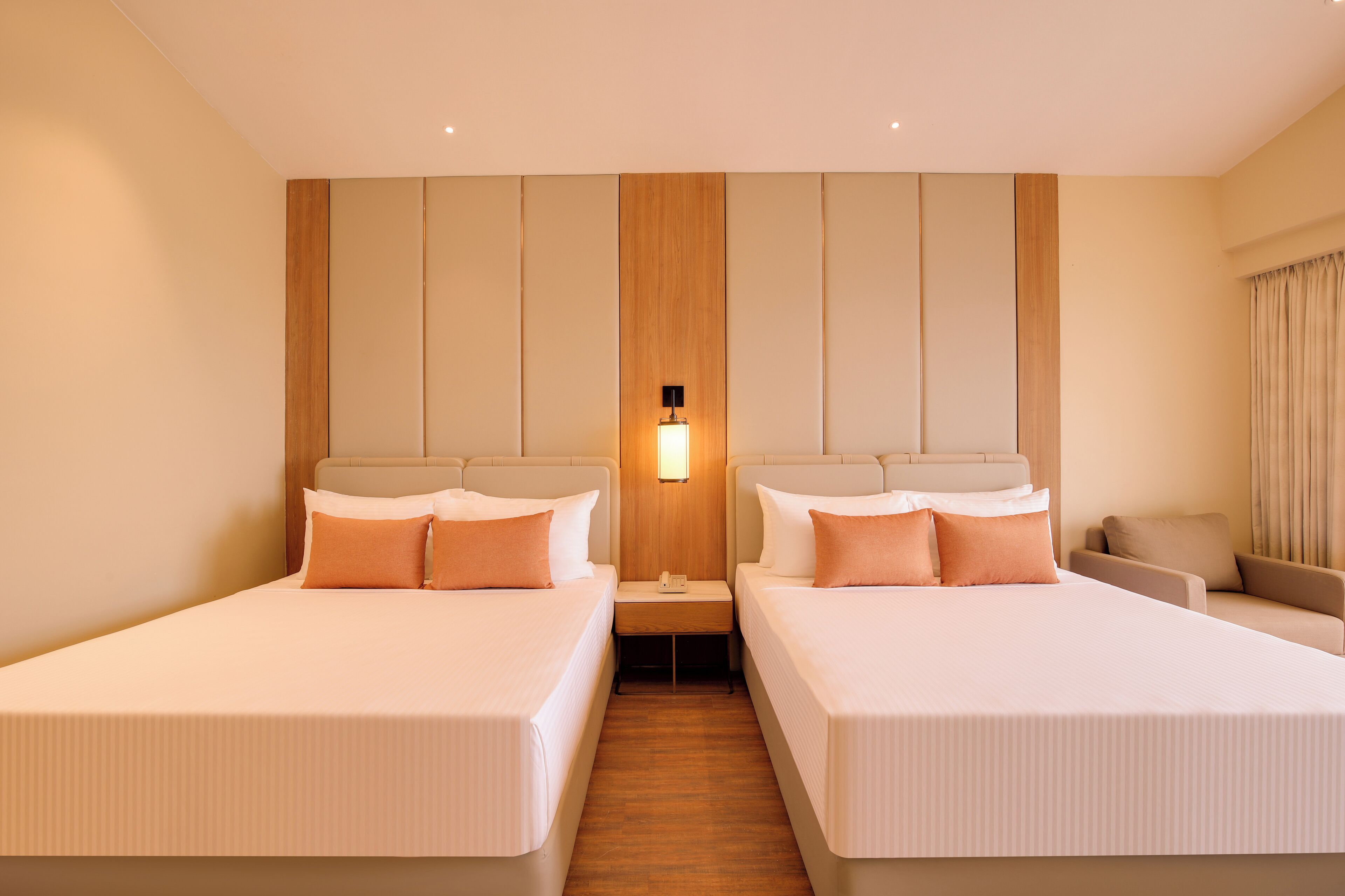A hotel room with two beds, a nightstand, a lamp, and a sofa.
