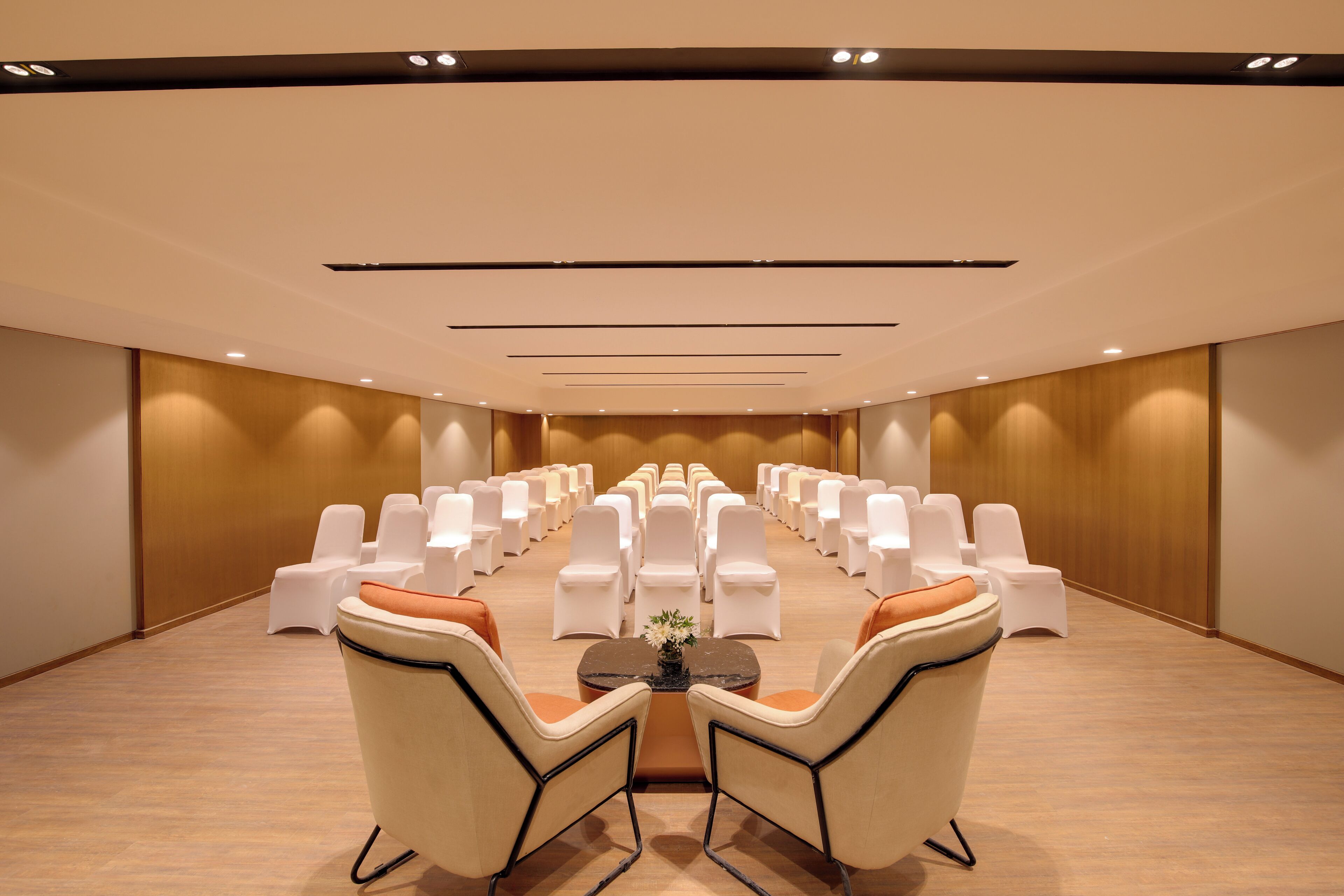 Un salón de conferencias con filas de sillas blancas y algunos sofás orientados hacia una mesa central con un jarrón de flores.
