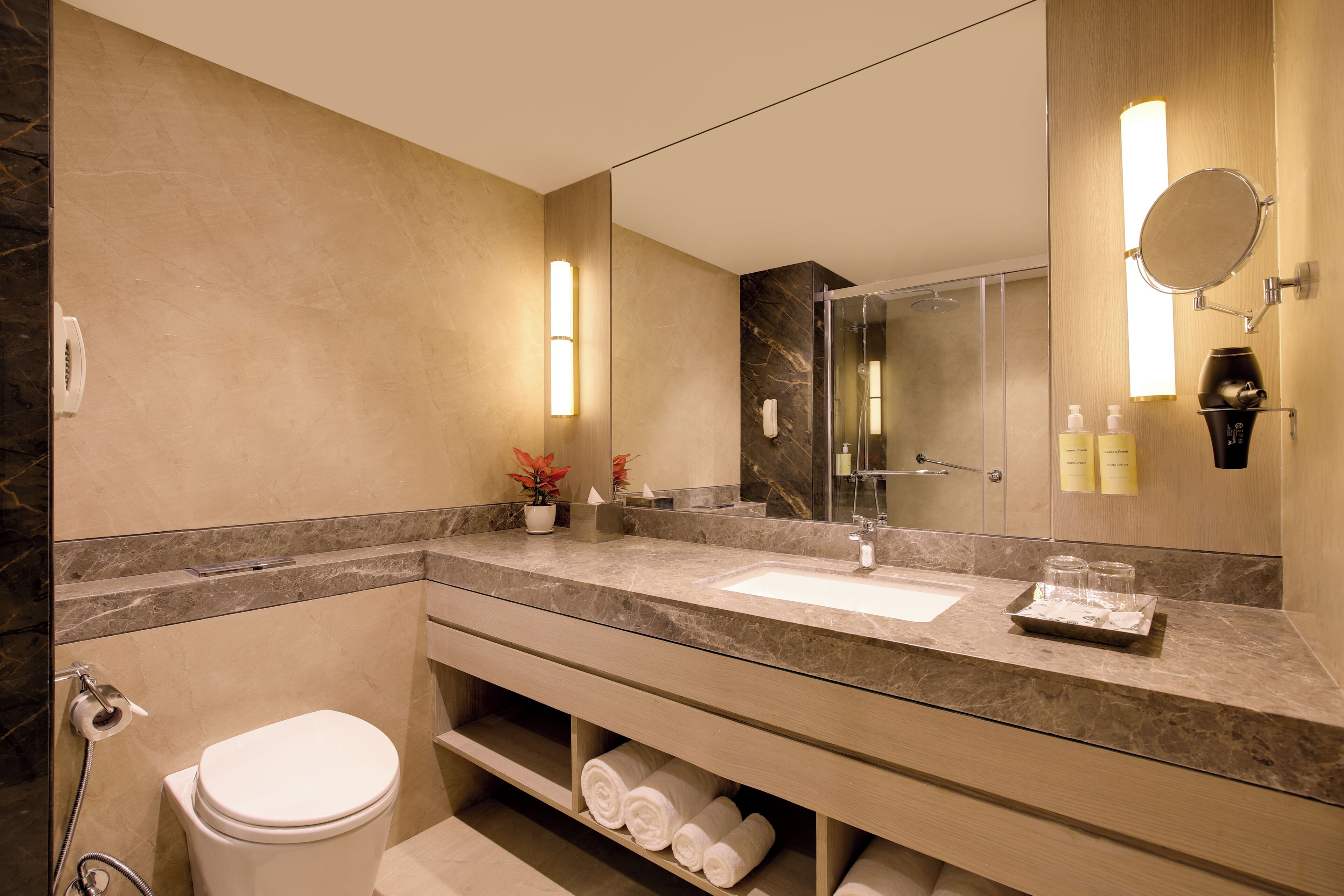 A modern bathroom with a marble vanity, a large mirror, a glass shower enclosure, and neatly rolled towels on a shelf.