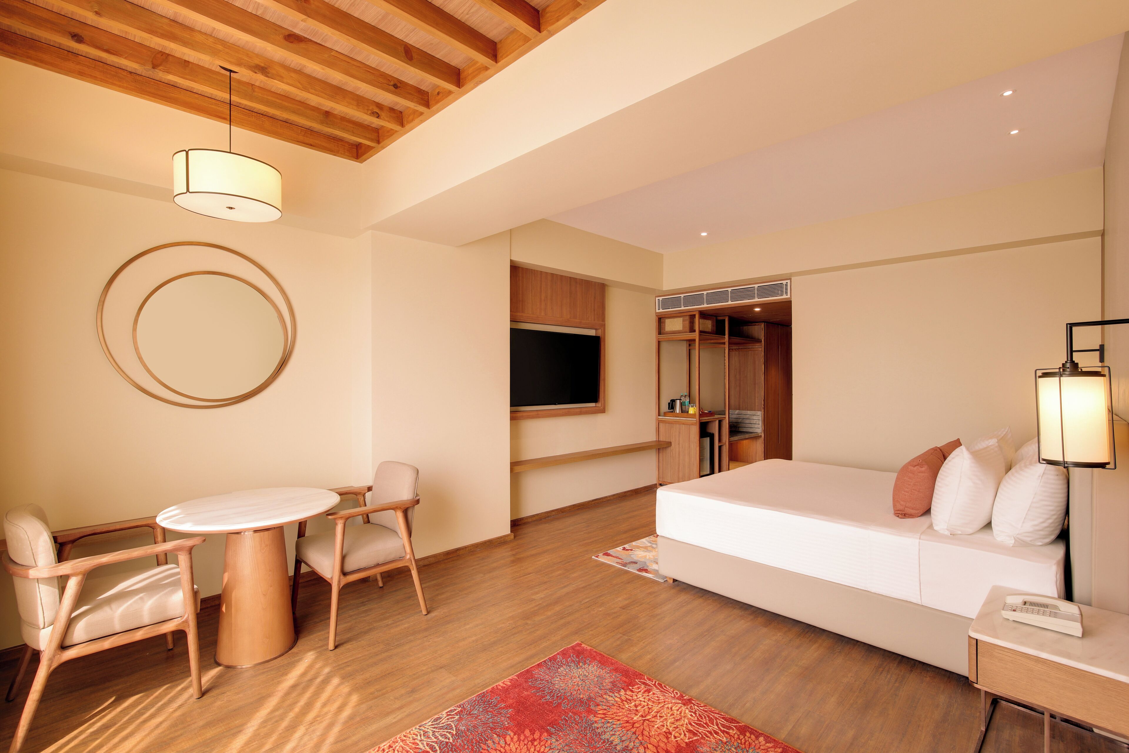 Habitación de hotel moderna con techo de madera, un espejo redondo, una cama con ropa blanca, una pequeña mesa con sillas y una alfombra roja con estampado.