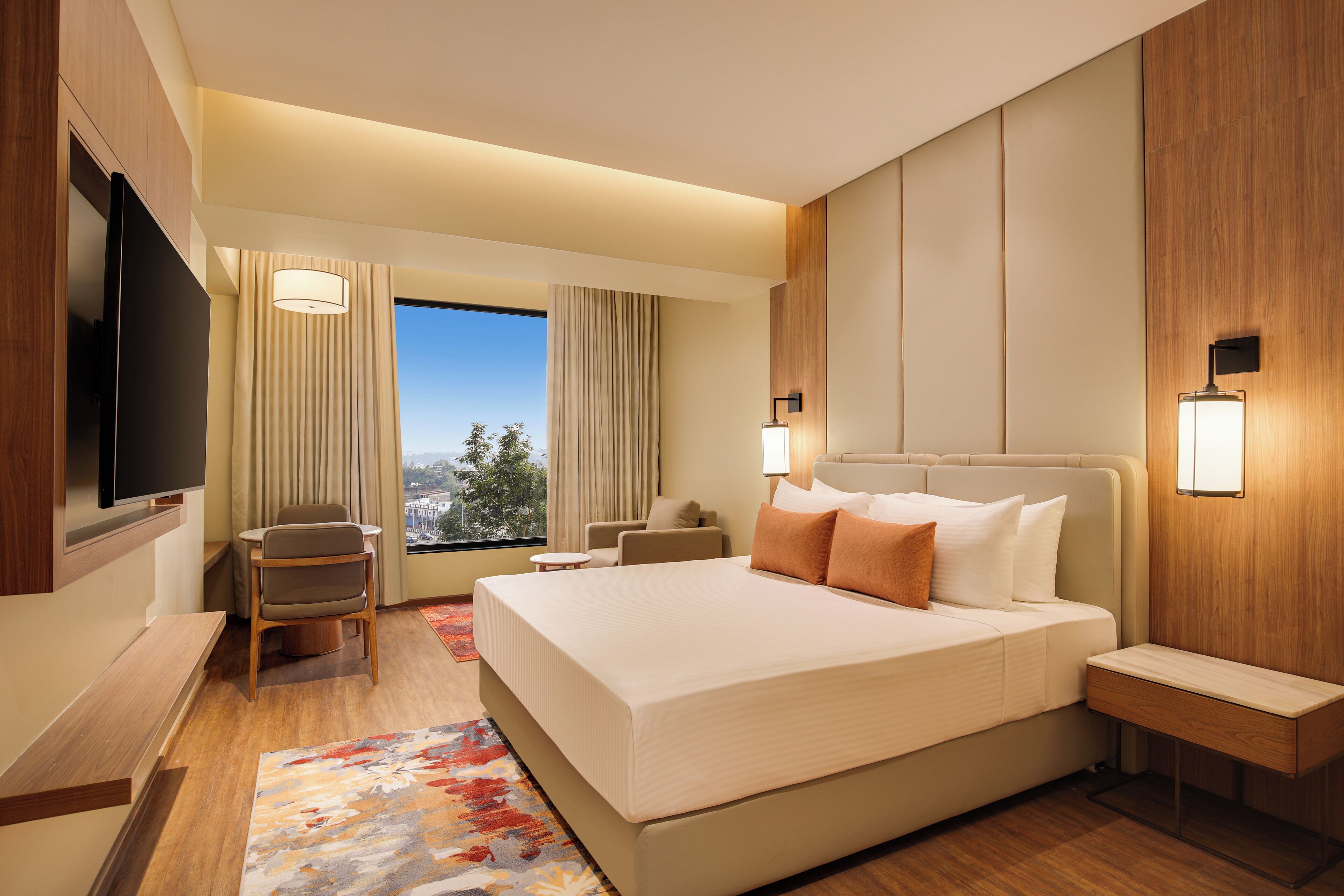 A hotel room with a large bed, a TV, a seating area with a chair and a small table, and a view of the outdoors.