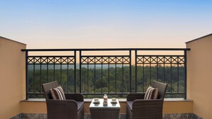 Een balkon met twee rieten stoelen, een klein tafeltje en uitzicht op een bebost gebied achter een metalen hekwerk.