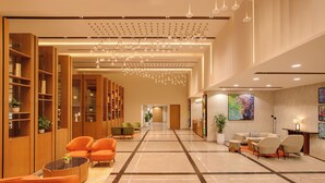 Een ruime lobby met een hoge plafond, moderne verlichting en een centraal zithoek met oranje stoelen.