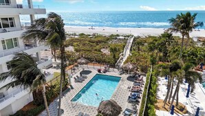 A resort with a swimming pool, lounge chairs, palm trees, and a view of the beach and ocean.