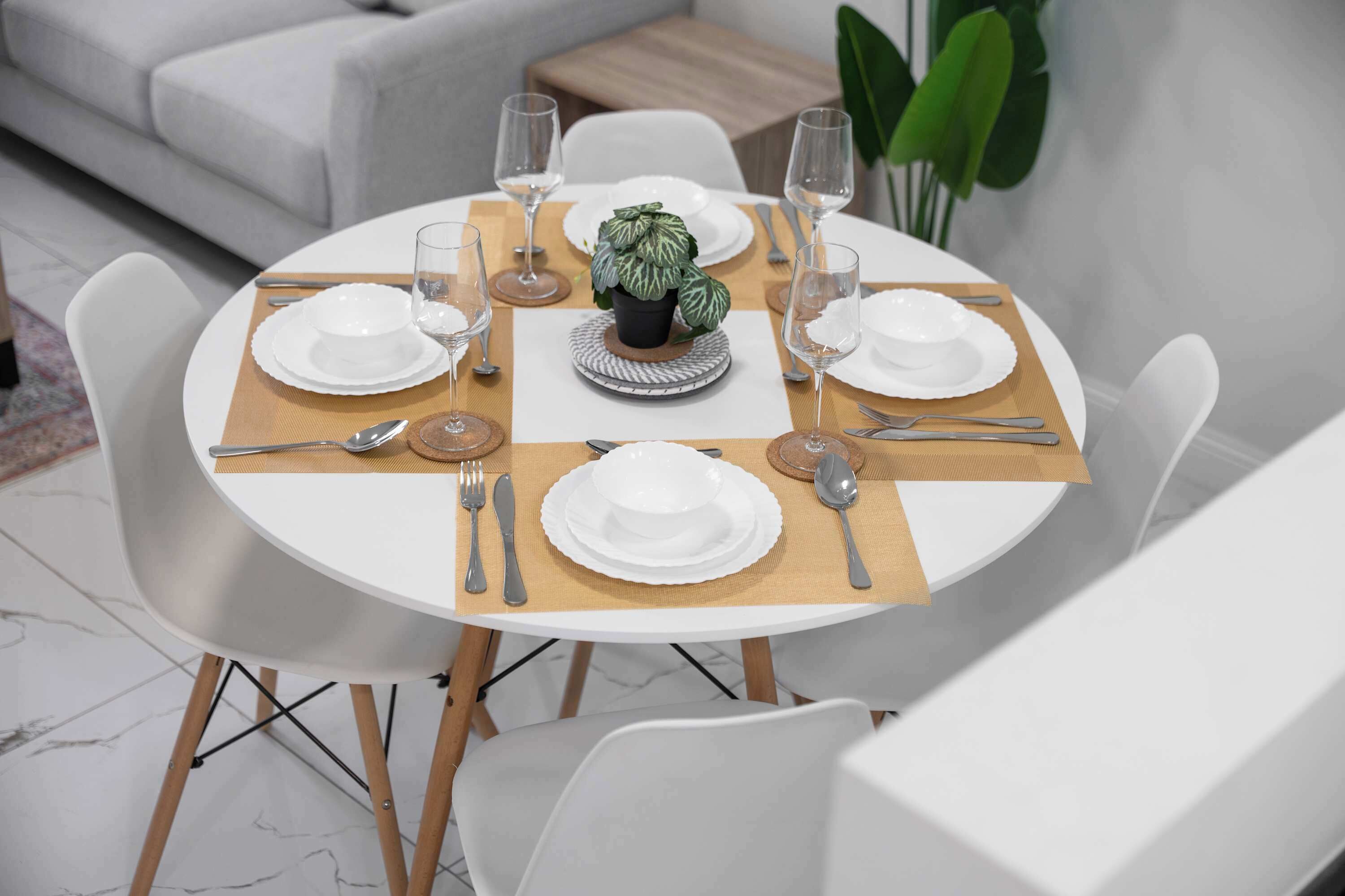 A round dining table with white plates, silverware, and wine glasses, surrounded by white chairs.